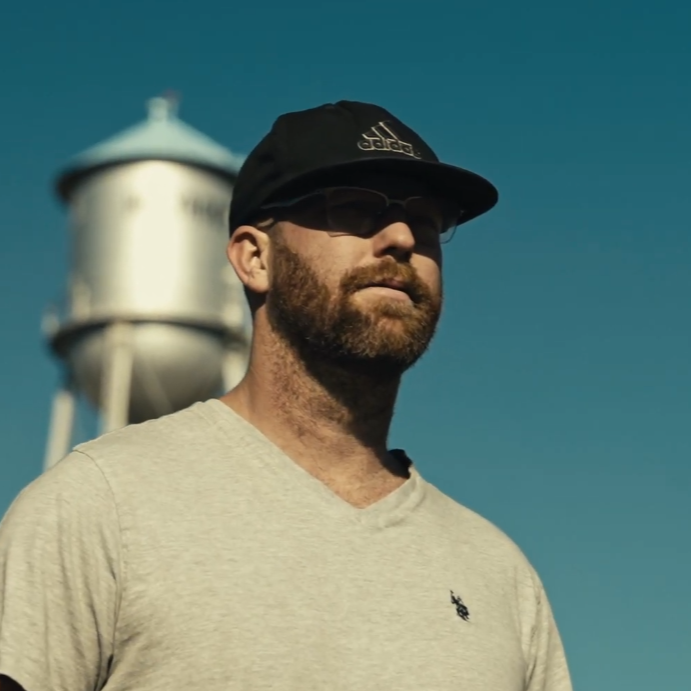 A person with a beard wearing a cap and sunglasses stands outdoors against a blue sky. A water tower is visible in the blurred background.