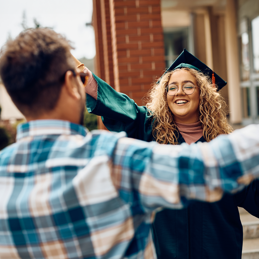A young woman in a graduation gown and cap smiles with open arms as she approaches a man in a plaid shirt, likely to embrace, outside a brick building.