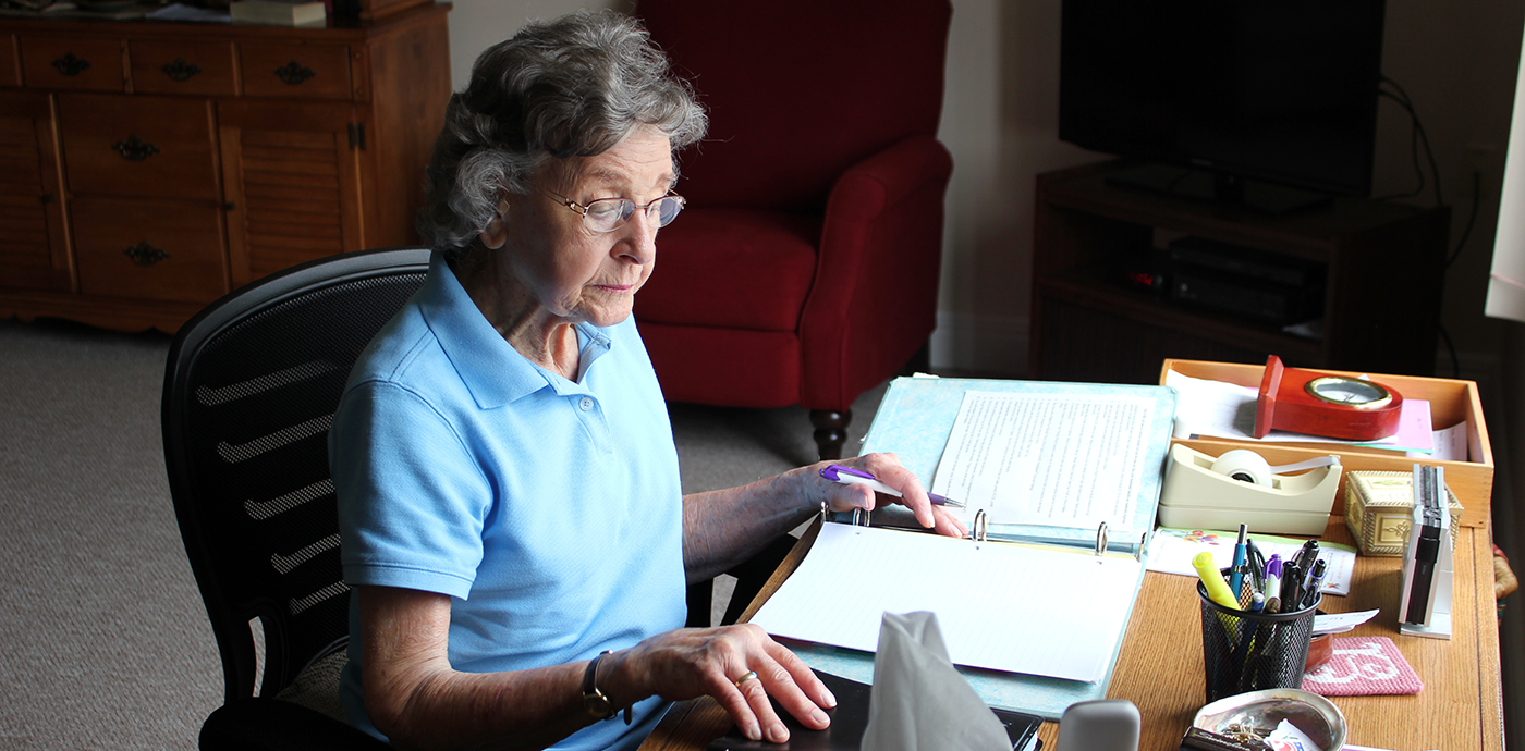 Elderly woman with glasses, wearing a blue polo shirt, sits at a desk with a binder open, reading a document. The desk holds various items including pens, a tissue box, and office supplies. A wooden cabinet and red armchair are visible in the background.