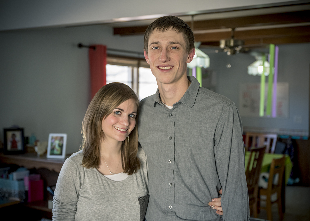 A man and woman stand together in a warmly lit living room, smiling at the camera. The man has short brown hair and is wearing a gray button-up shirt. The woman has long light brown hair and is wearing a gray long-sleeve shirt. The room has light blue walls and a large window.