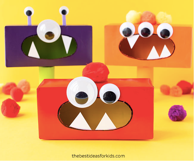 Three colorful monster-themed tissue box crafts are displayed against a yellow background. The boxes are decorated with large googly eyes, sharp white teeth, and small pom-poms. The monsters are in purple, orange, and red colors.