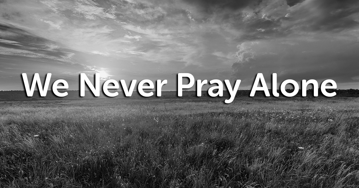 A black-and-white image shows a field of tall grass under a cloudy sky with the sun partially visible. The text "We Never Pray Alone" is superimposed over the image in bold white letters.