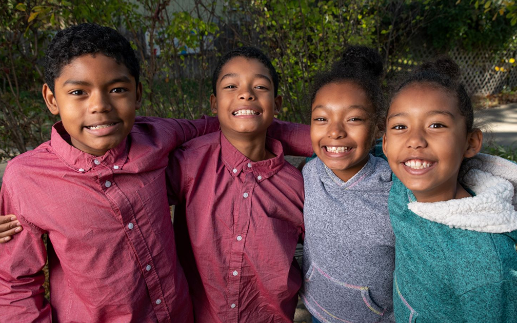 Four children stand closely together outdoors, smiling at the camera. The two boys on the left wear red shirts, while the two girls on the right wear gray and teal tops. Trees and foliage are visible in the background.