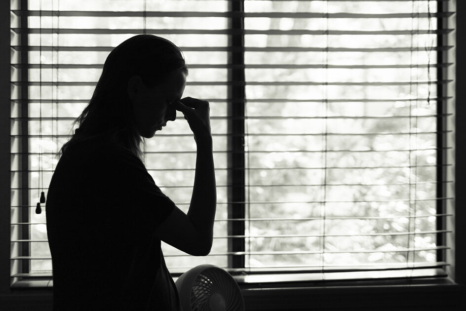 A person stands in front of a window with closed blinds, holding their fingers to their forehead. The scene is backlit, creating a silhouette effect. Sunlight filters through the blinds, casting a diffused light into the room.