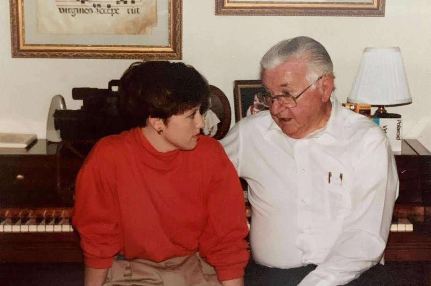 An older man and a younger woman sit together in front of a piano, engaged in conversation. The man wears a white shirt and glasses, while the woman is dressed in a red sweater. Framed pictures and a lamp on a table are visible in the background.