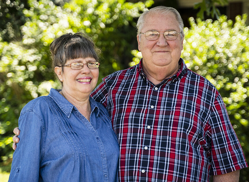 An older couple standing side by side outdoors, both smiling. They are wearing glasses, and the woman is dressed in a blue shirt while the man is wearing a red and blue plaid shirt. The background is green foliage.