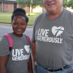 Two people stand side by side, both wearing gray "Live Generously" T-shirts. The person on the left has a pink backpack and smiles at the camera. The person on the right wears glasses, a hat, and also smiles. Trees and a building are visible in the background.