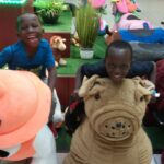 Two children are sitting on plush animal ride-on toys in a play area. The child on the left is on an orange toy while the one on the right is on a brown toy. Both children are smiling and appear to be having fun. Other plush animals are visible in the background.