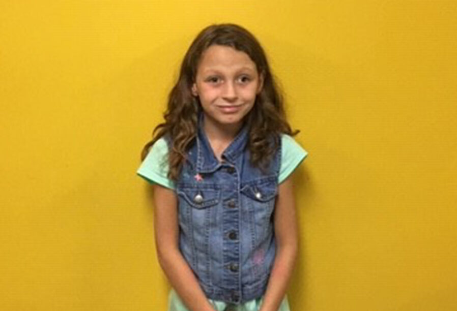 A young girl with long brown hair stands against a yellow background. She is wearing a light green short-sleeved top and a sleeveless denim vest. She is looking at the camera with a neutral expression.