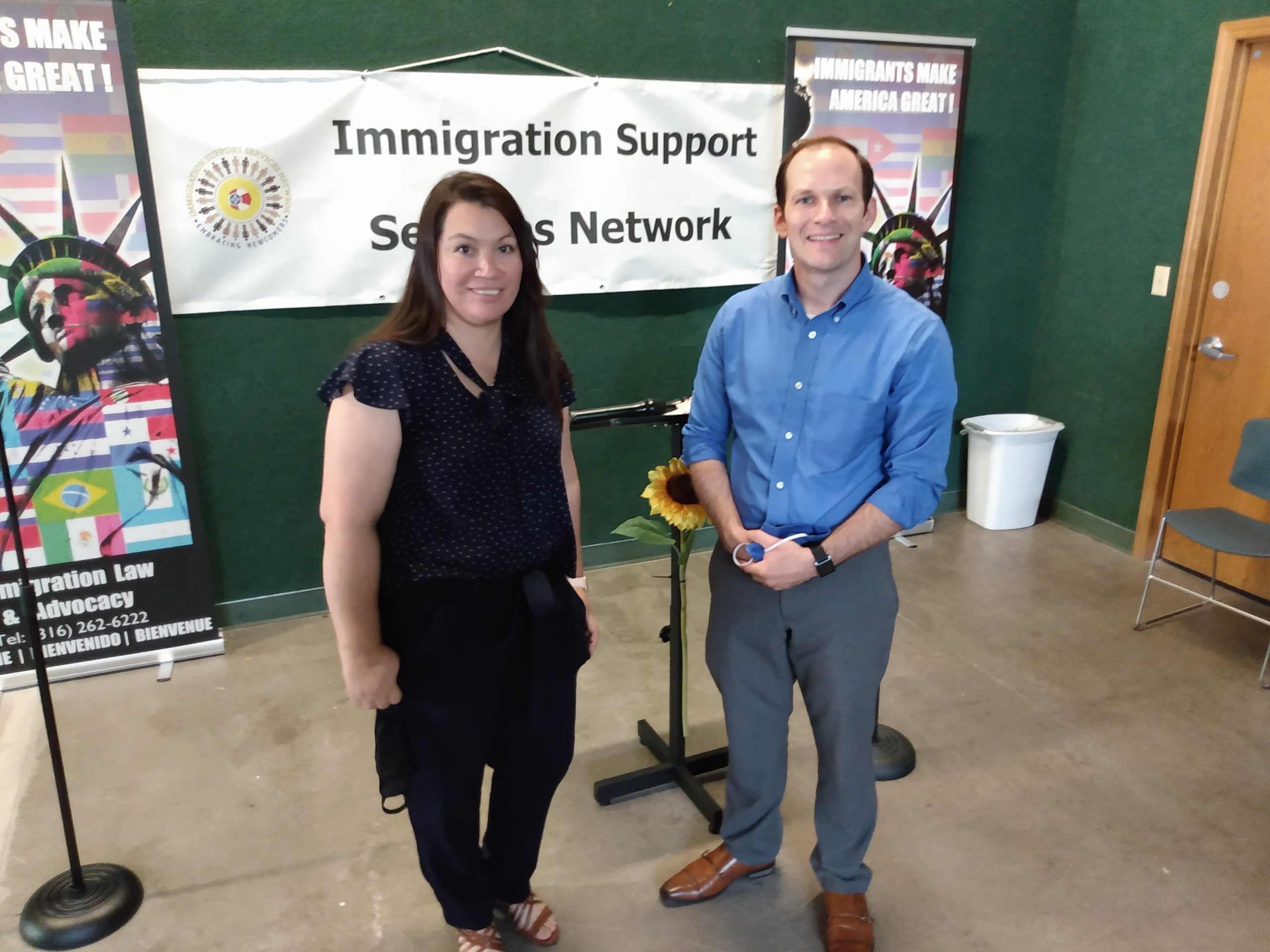 Two individuals stand in front of a banner that reads "Immigration Support Services Network." The woman on the left is wearing a dark blouse and pants, while the man on the right is dressed in a blue shirt and gray pants. A small table with a sunflower is nearby.
