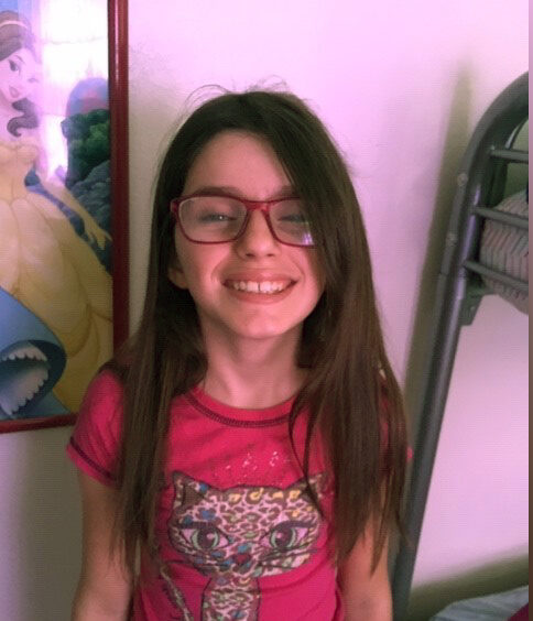 A young girl wearing red glasses and a pink shirt with a cat graphic smiles at the camera. She has long brown hair. A framed picture of a cartoon character in a yellow dress is partially visible on the wall behind her.