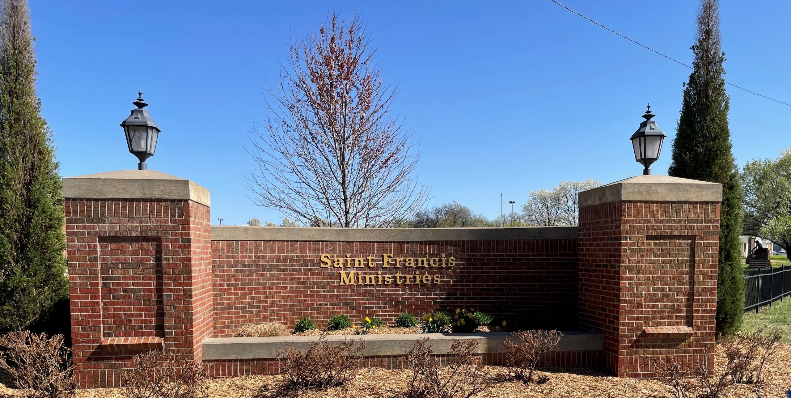 The image shows a red brick sign for Saint Francis Ministries. The sign is flanked by two brick columns topped with black lamp posts. A tree and some plants are in the background, and the sky is clear and blue. Shrubs are planted at the base of the sign.