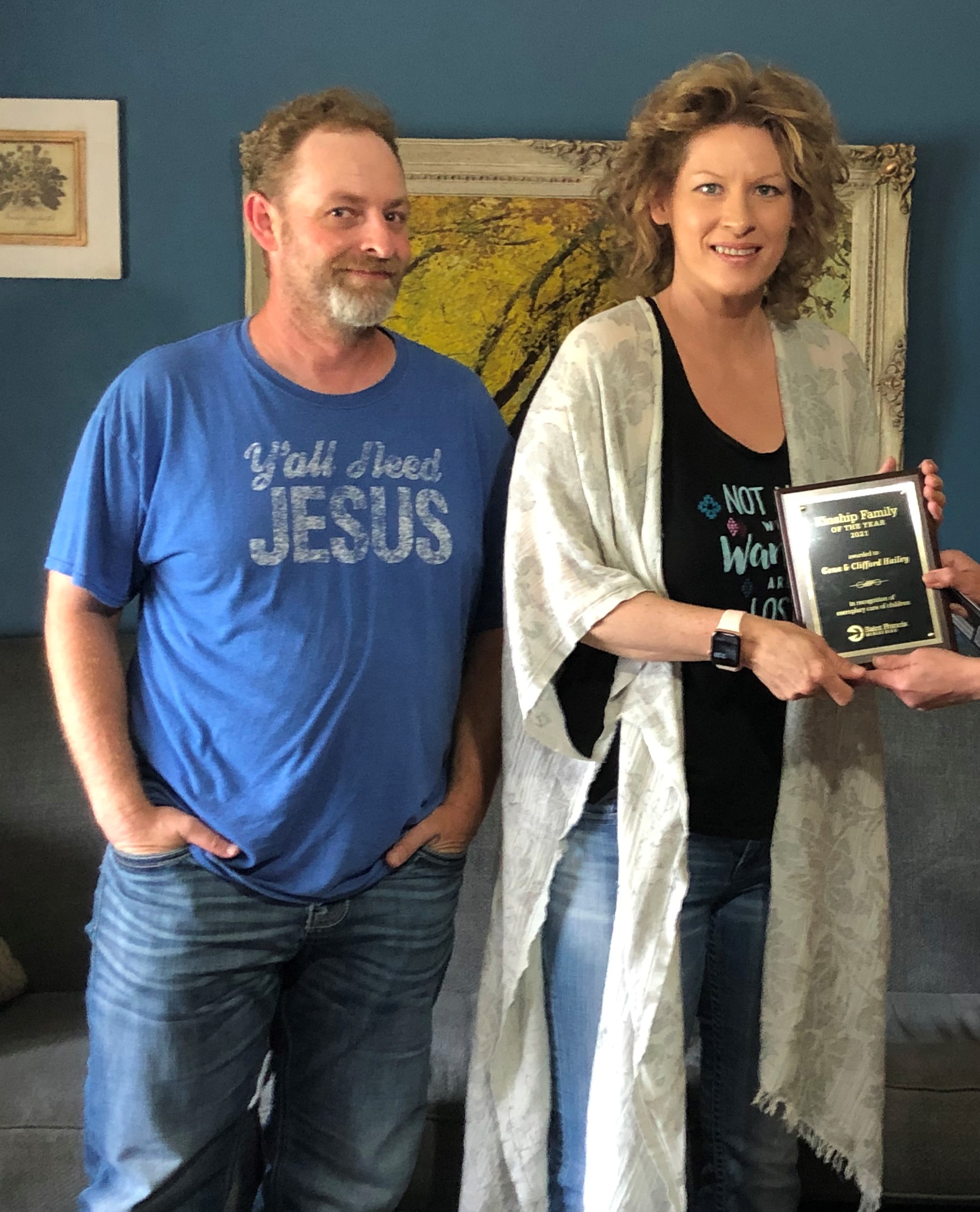 A man and a woman stand side by side indoors. The man is wearing a blue t-shirt that reads "Y'all Need Jesus." The woman, holding a plaque, is wearing a black top and a light cardigan. They are both posing in front of a dark blue wall with framed art.