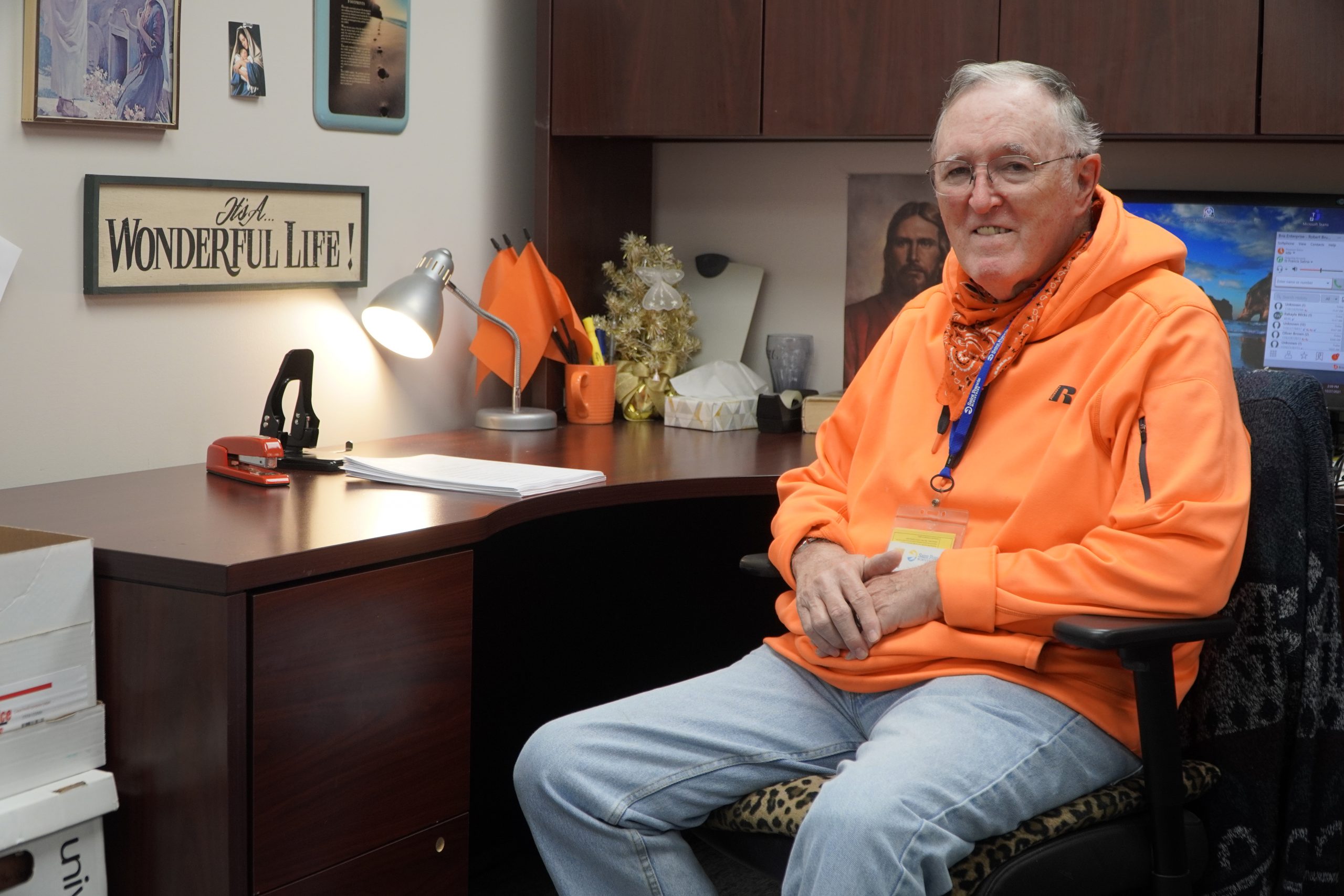 An elderly man wearing an orange hoodie sits in an office chair beside a polished wooden desk. The desk has a lamp, office supplies, and a "It's a Wonderful Life!" sign on the wall. A computer monitor and personal items are visible in the background.
