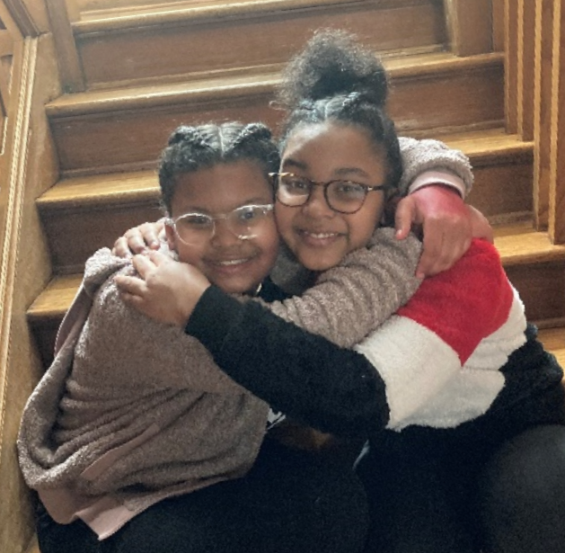 Two children sit on wooden stairs and embrace each other with smiles on their faces. They both wear eyeglasses and casual clothing. One wears a red, white, and black sweater, while the other wears a brown jacket over a black shirt.