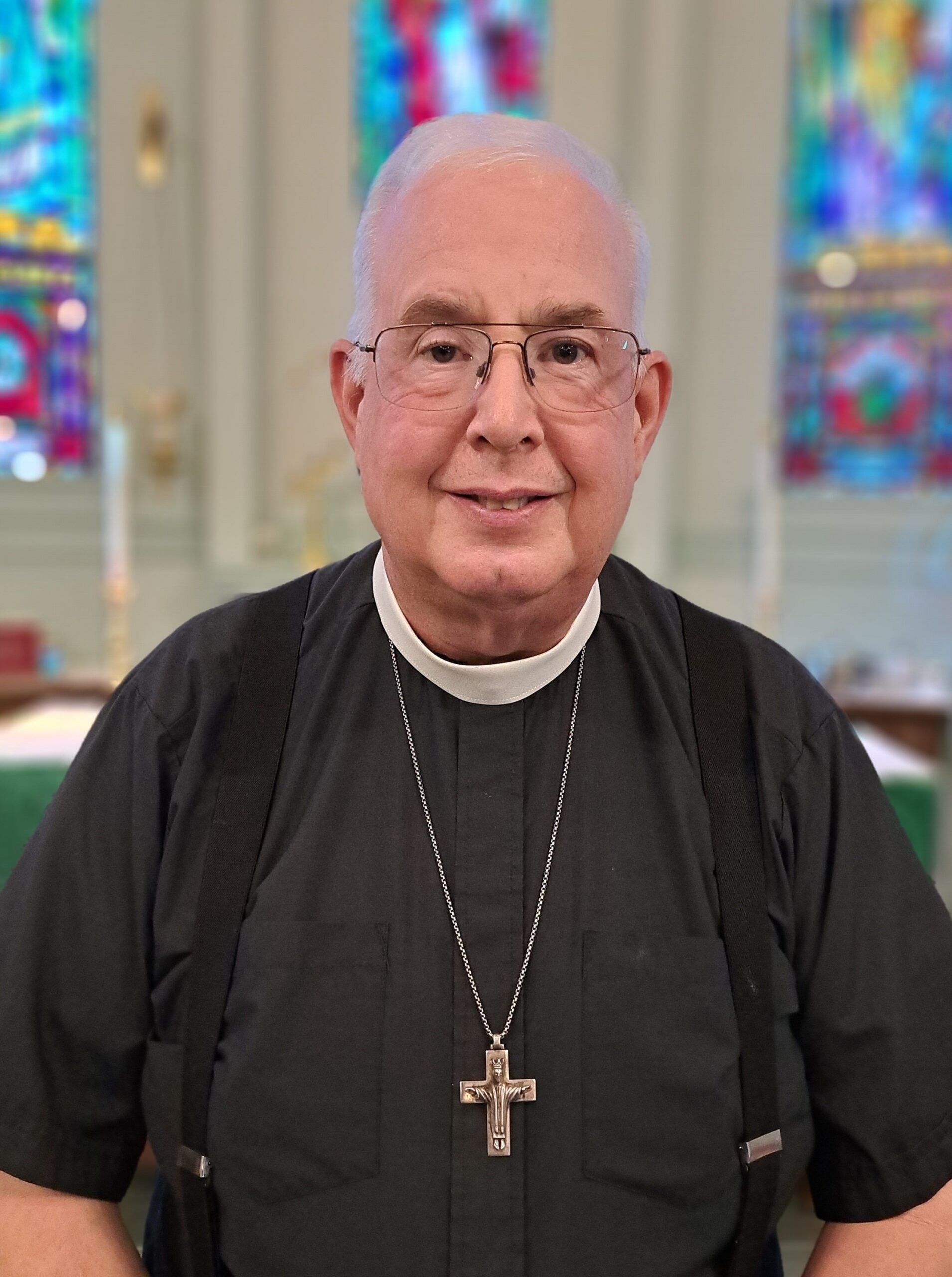 A man wearing a clerical collar, black shirt, and glasses stands in a church with colorful stained glass windows in the background. He has white hair and is smiling. A cross necklace hangs around his neck.