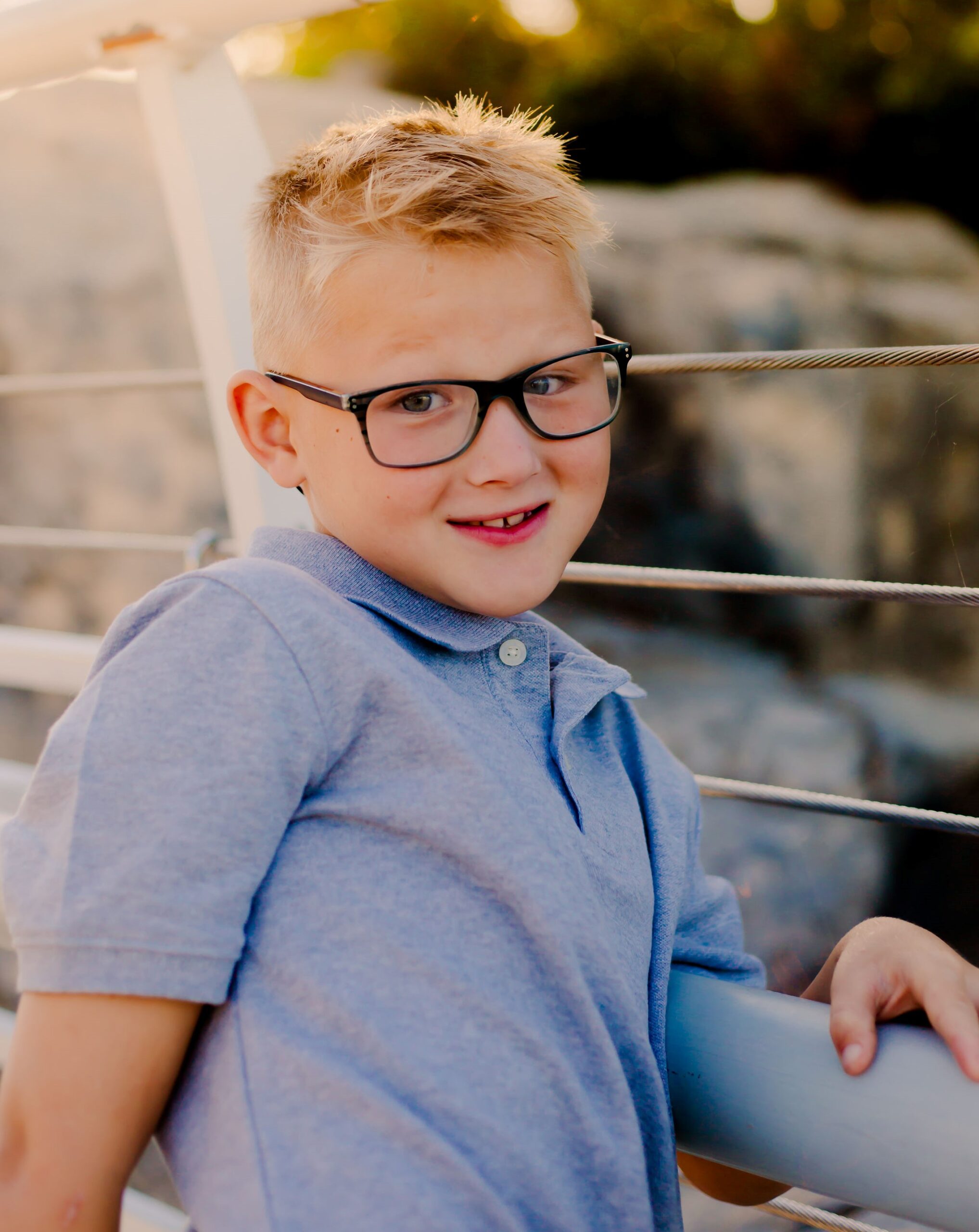 A young boy with short blond hair and glasses stands against a railing outdoors. He is smiling slightly and wearing a light blue polo shirt. The background includes a blurry natural setting with rocks and greenery.