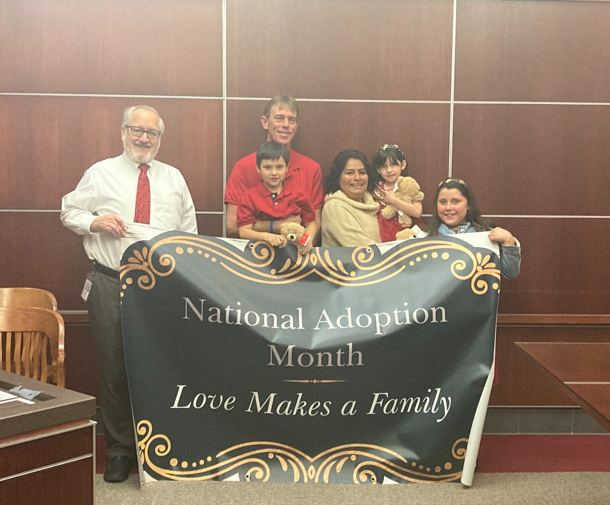 A group of six people, including two adults holding children, stand together in a courtroom. They are holding a large banner that reads "National Adoption Month: Love Makes a Family." The background features wooden paneling with some chairs and a table.