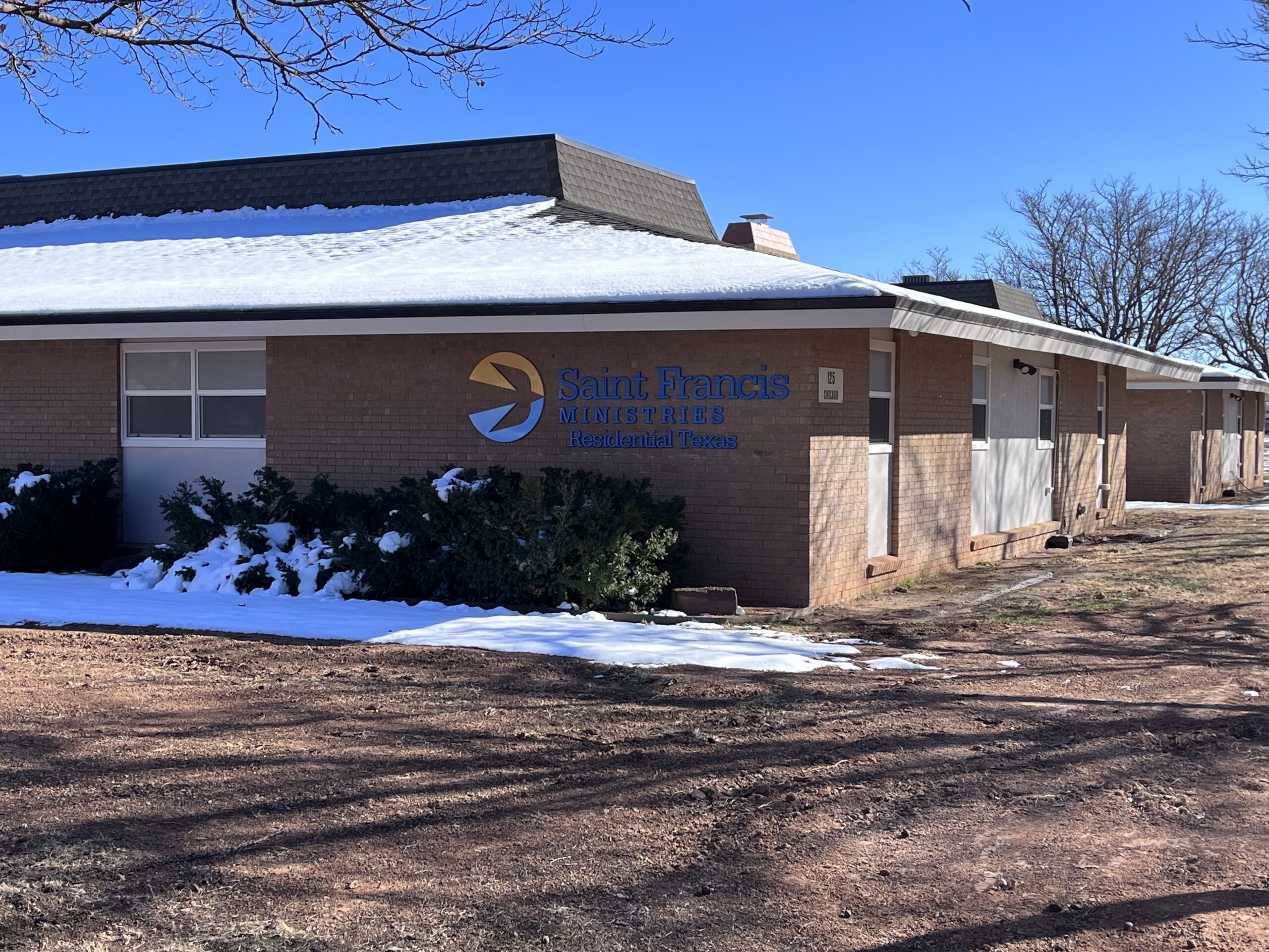 A one-story brick building with the sign "Saint Francis Ministries Residential Texas" on the front wall. Snow is on parts of the ground and bushes near the building. Trees without leaves are visible in the background under a clear blue sky.