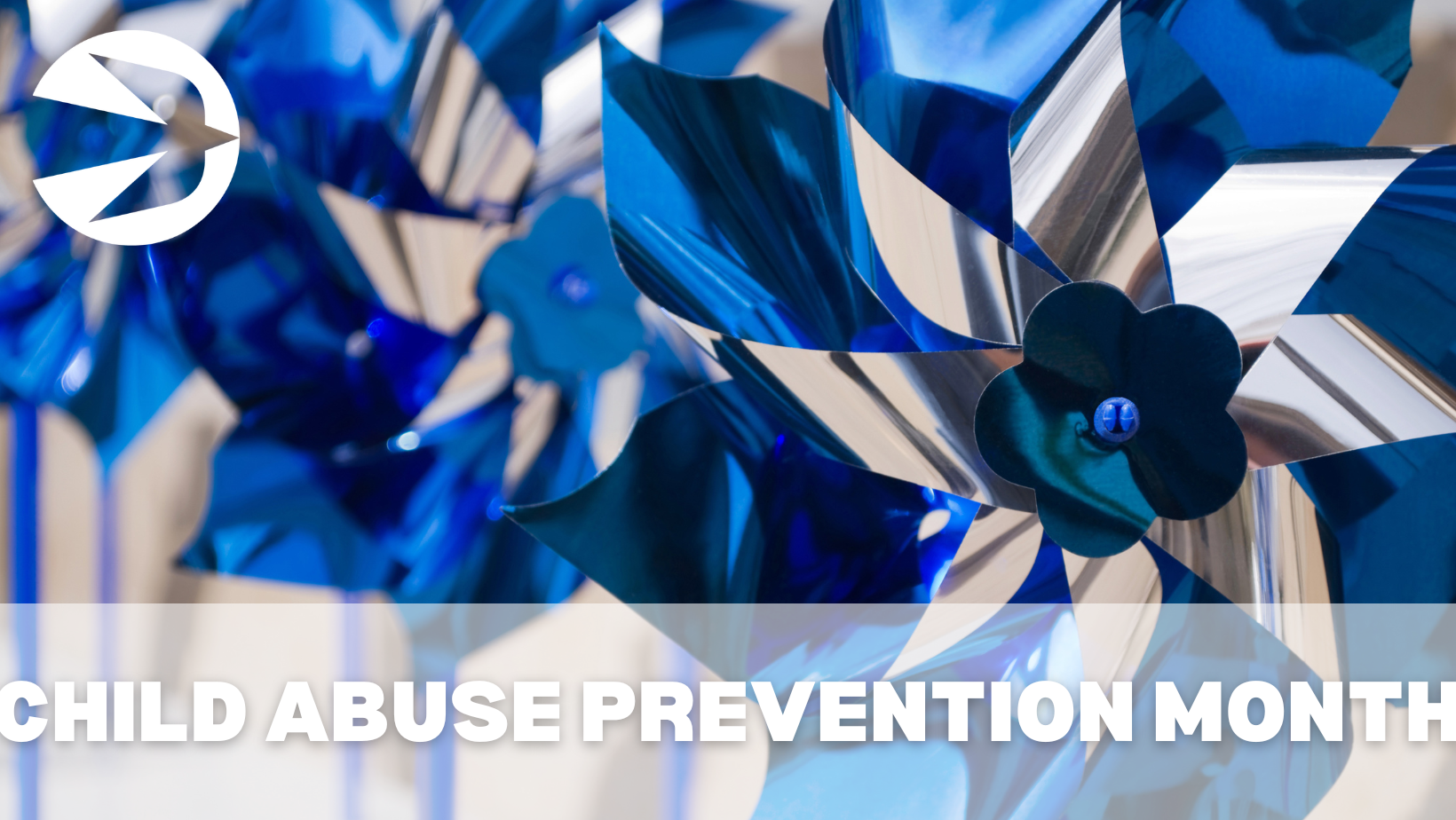 A close-up of blue and silver pinwheels with the text "CHILD ABUSE PREVENTION MONTH" at the bottom. A logo with a white arrow inside a blue circle is at the top left corner. The background is slightly blurred, highlighting the vibrant pinwheels.