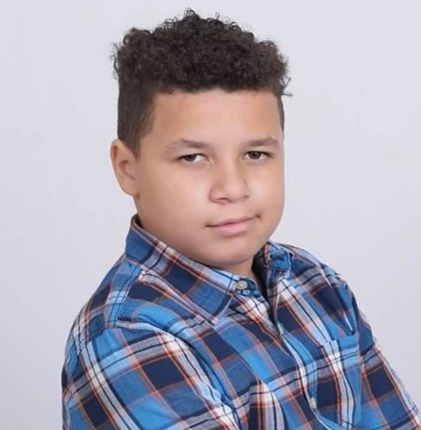 A young boy with short, curly hair is wearing a blue, white, and red checkered shirt and looking at the camera with a neutral expression. The background is plain white.