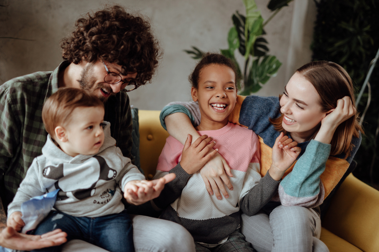 A joyful family sitting together on a couch with parents playing with their two children featuring a toddler and an older girl, in a cozy living room.