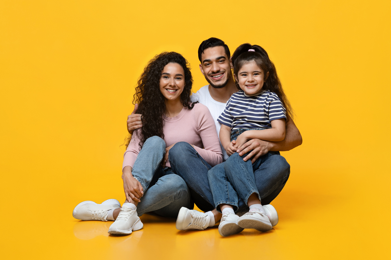 A happy family consisting of a mother, father, and young daughter sitting together, smiling at the camera against a vibrant yellow background. All are wearing casual clothes and white sneakers.