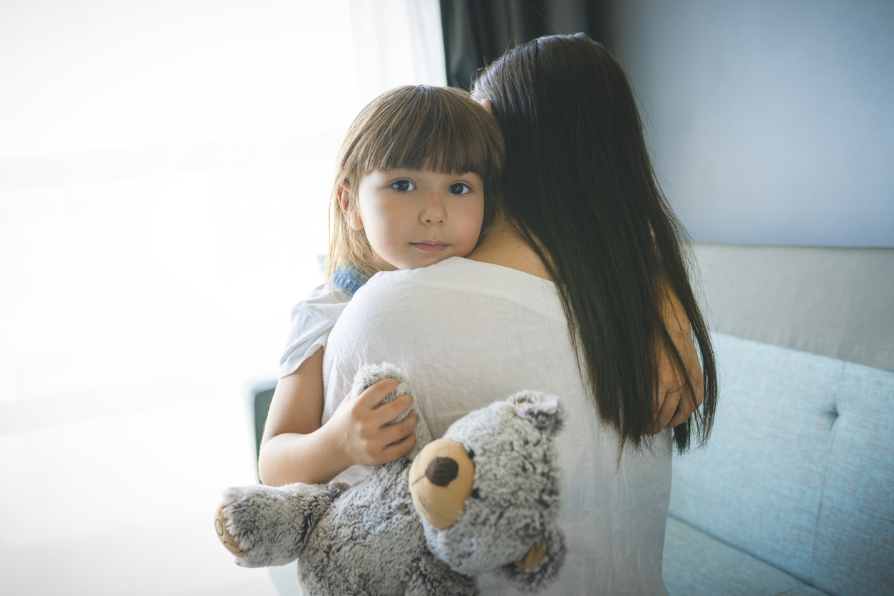 A young girl with a teddy bear hugged closely by her mother, only the mother's back is visible. They are sitting on a couch in a brightly lit room.