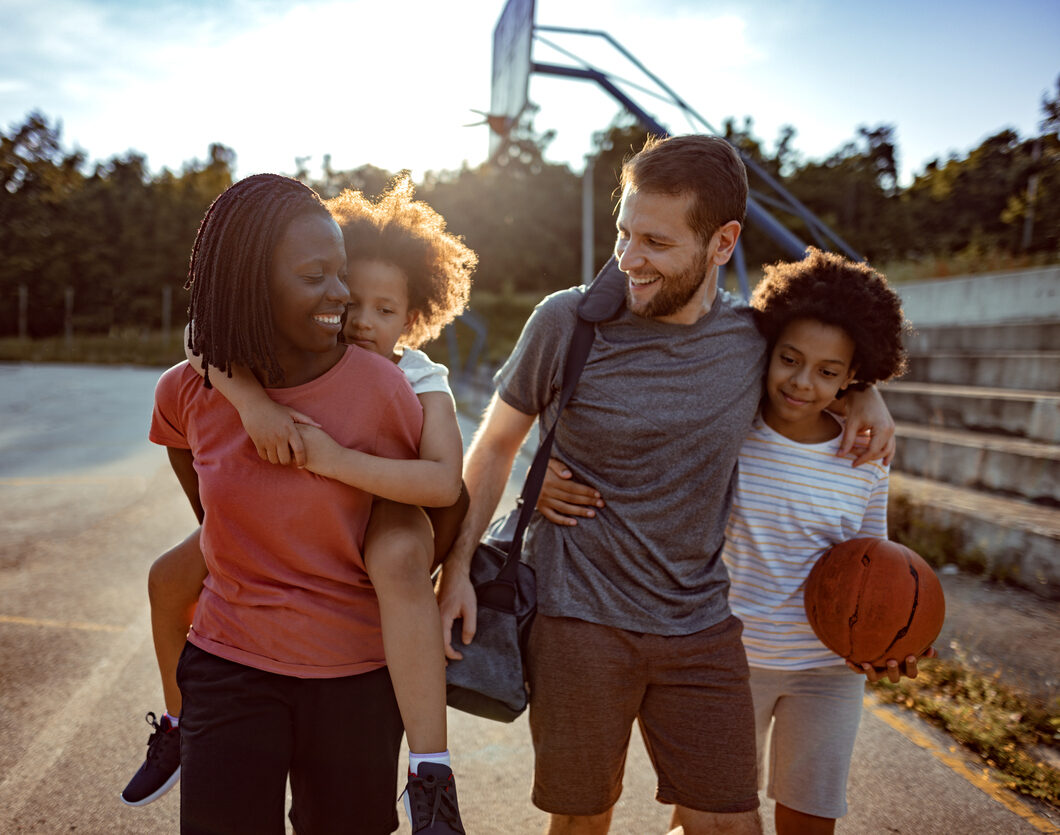 A joyful family of four walking in a sunlit park, with a young child piggybacking on the father's back and another holding a basketball, engaging playfully.