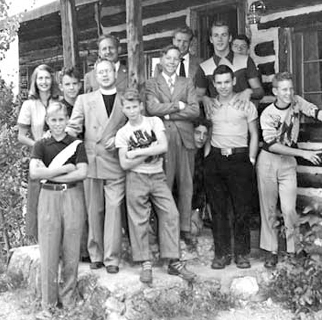 A group of thirteen people, consisting of adults and children posing in front of a rustic cabin with some standing, others sitting, in casual attire, exuding a warm