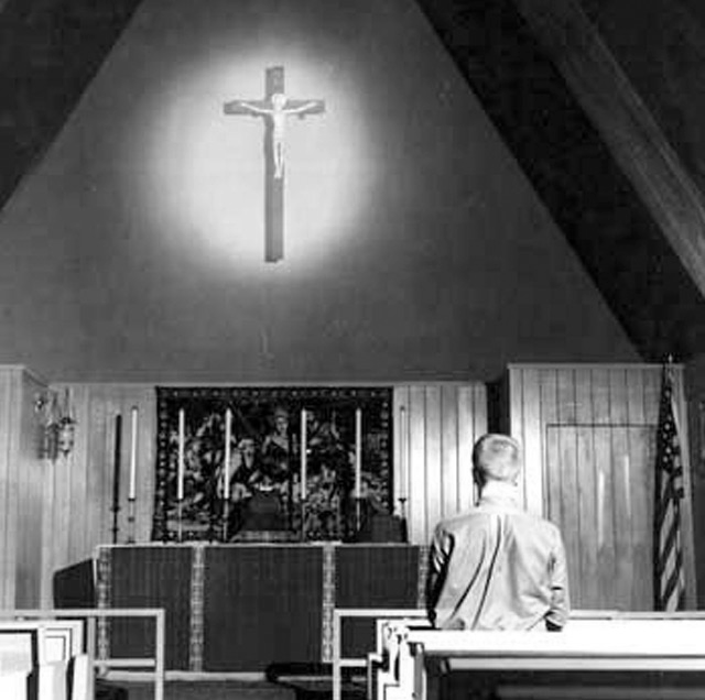 A man sits alone in a church pew, looking towards an illuminated crucifix above the altar. The church has a triangular ceiling and simple wooden walls.