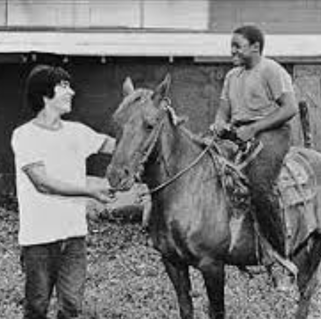 Black and white photo of two people with a horse: one in a white t-shirt holding the horse's reins, the other in a tank top smiling and looking at the first person.