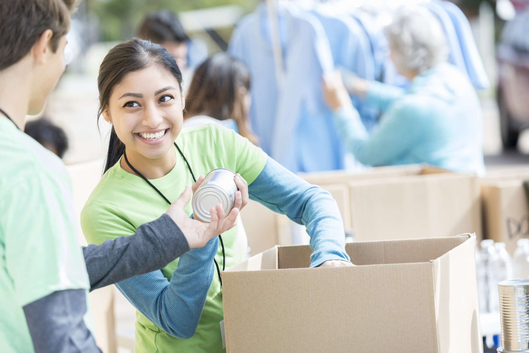 A woman in a green shirt smiles as she hands a canned good to a person. They are outdoors, and there are cardboard boxes and people in the background. The scene appears to be a volunteer event or food drive.