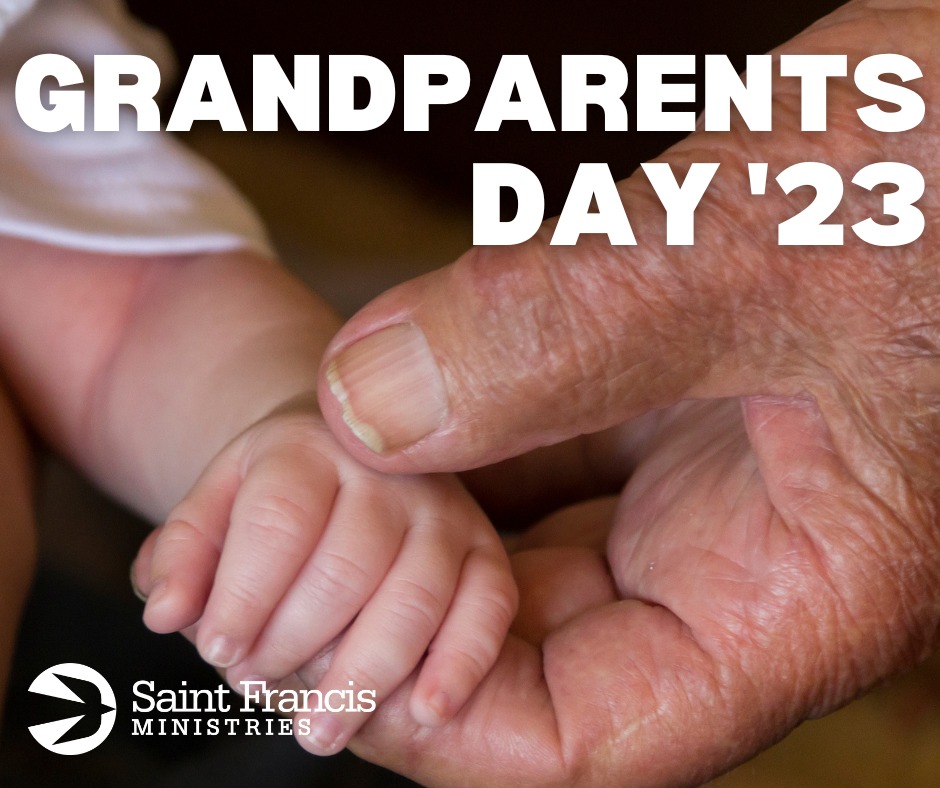 A close-up image shows an elderly hand holding a baby's hand. The text "GRANDPARENTS DAY '23" is written at the top of the image. The bottom left corner features the Saint Francis Ministries logo and name.
