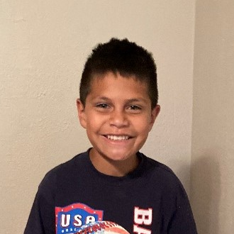 A young boy with short dark hair smiles at the camera. He is wearing a dark blue T-shirt with a USA baseball logo on it. He is holding a small orange and white basketball. He is standing in front of a plain, light-colored wall.