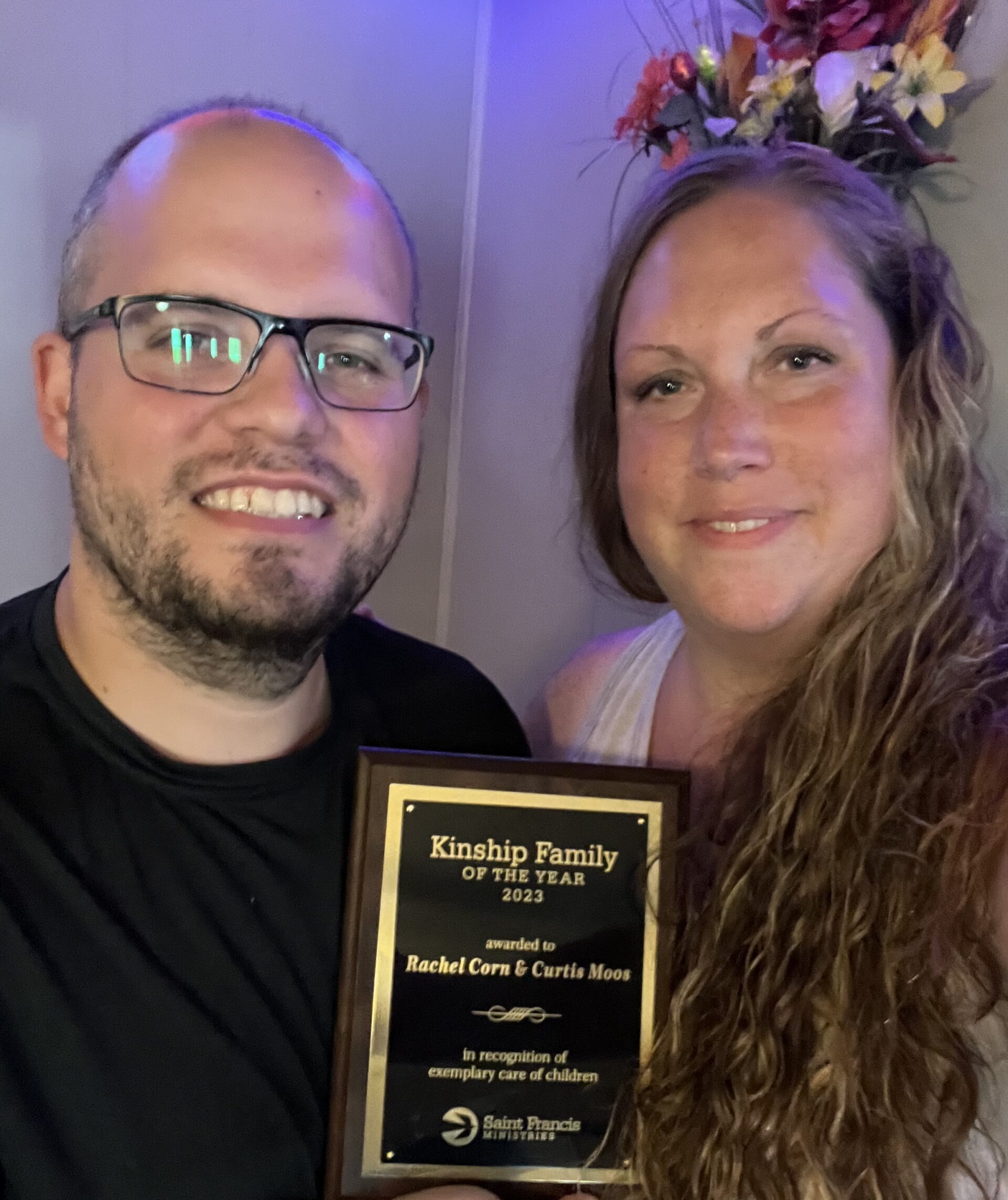 A man and a woman are smiling while holding a plaque that reads, "Kinship Family of the Year 2023 awarded to Rachel Corn & Curtis Moos in recognition of exemplary care of children." They stand in front of a flower arrangement.
