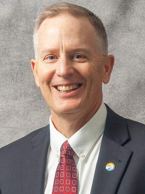 A man with short gray hair is smiling and wearing a dark suit, white shirt, and a red patterned tie. He also has a round pin with a multicolored logo on his suit lapel. The background is a plain gray texture.