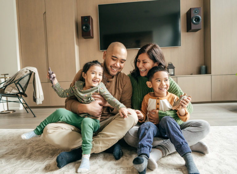 A family of four sits together on the floor of a living room, smiling and laughing. The children, one boy and one girl, sit on their parents' laps. In the background, there is a wall-mounted television and two speakers.