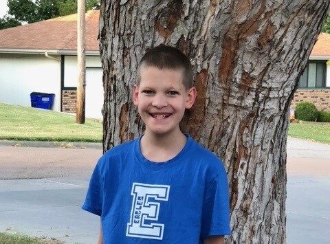 A boy with short hair smiles while standing outdoors in front of a tree. He is wearing a blue T-shirt with the word "Eagles" printed on it. There are houses in the background and a blue trash can visible on the left.