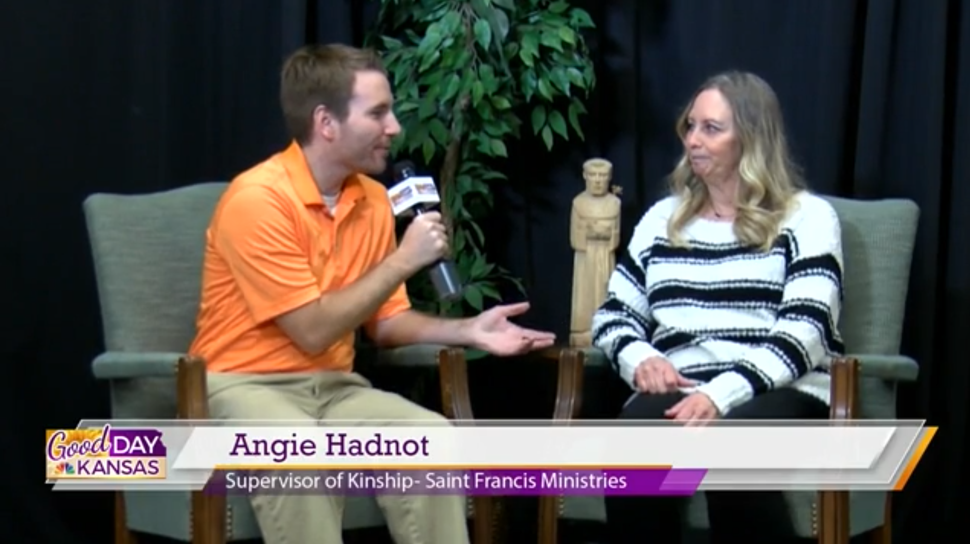A man in an orange polo shirt is interviewing a woman in a black and white striped sweater. They are seated in chairs on a stage with a small statue and a plant in the background. The screen text reads "Good Day Kansas," "Angie Hadnot," and "Supervisor of Kinship - Saint Francis Ministries.