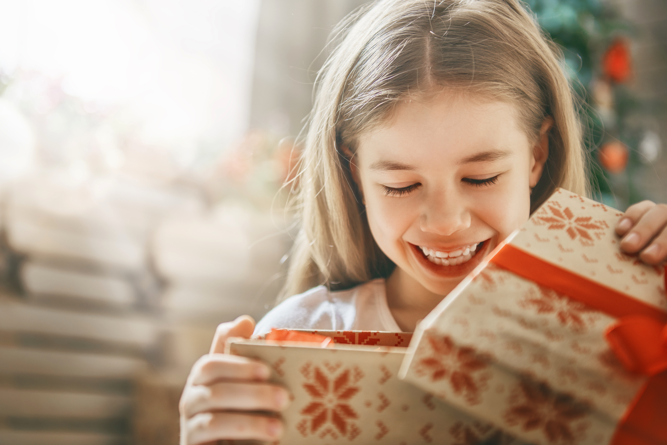A young girl with long hair is smiling and looking down at an open gift box wrapped in festive paper with red snowflake patterns and a red ribbon. The background appears to be decorated for the holiday season.
