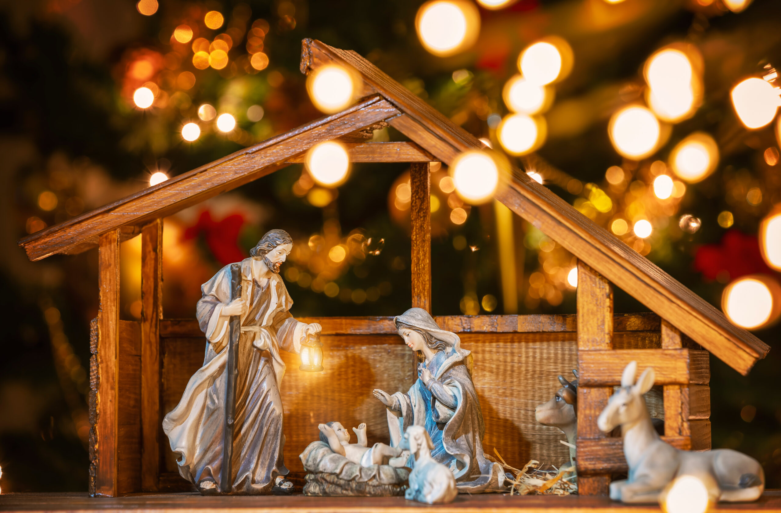 A wooden nativity scene featuring figurines of Joseph holding a lantern, Mary kneeling, baby Jesus in a manger, and animals. The background is adorned with festive Christmas lights.