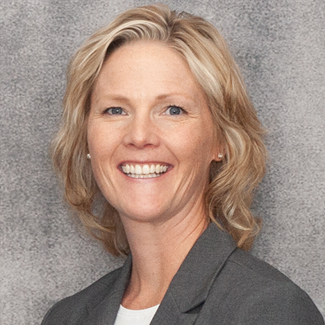 A woman with shoulder-length blonde hair smiles at the camera. She is wearing a gray blazer over a white top. The background is a gray, textured surface.