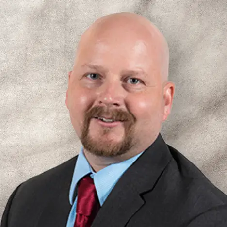 A bald man with a light complexion smiles softly in a professional portrait. He has a goatee and is wearing a dark suit jacket, light blue dress shirt, and a red tie. The background is a textured, light-colored surface.