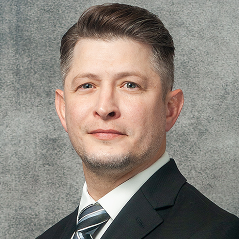 A man with short, neatly combed hair is wearing a black suit, white shirt, and a striped tie. He is posing in front of a textured grey background, looking directly at the camera with a neutral expression.