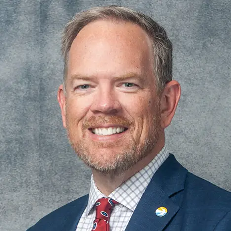 A man with short light hair and a beard is smiling. He is wearing a dark blue blazer, a checkered shirt, and a red patterned tie. There is a round pin on his blazer lapel. The background is a textured gray surface.