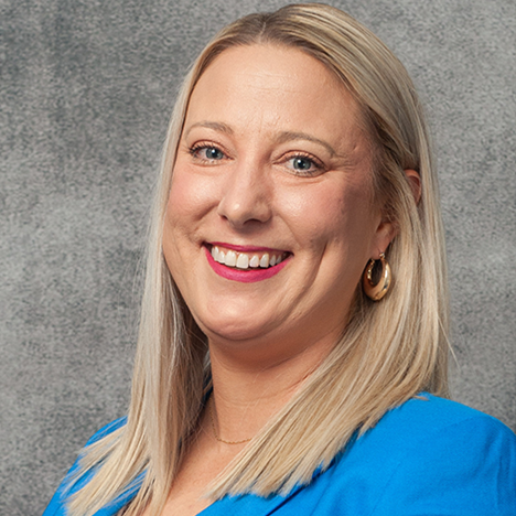 A woman with straight, shoulder-length blond hair is smiling and facing the camera. She is wearing a blue blazer, gold hoop earrings, and a gold necklace. The background is a textured gray surface.