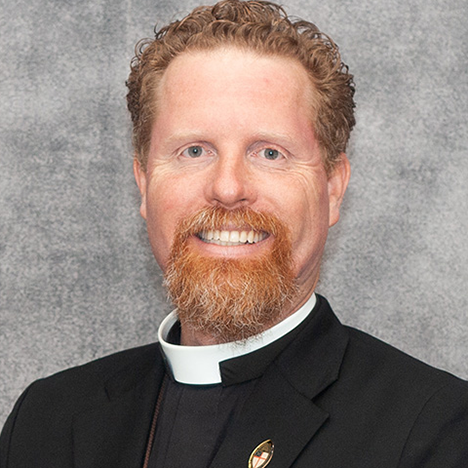 A man with curly red hair and a beard is smiling at the camera. He is wearing a clerical collar and a black blazer with a circular pin featuring a shield and cross on the lapel. The background is a plain, textured gray.
