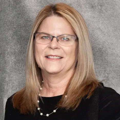 A middle-aged woman with straight, shoulder-length blonde hair is smiling at the camera. She is wearing glasses, a black top, and a pearl necklace. The background is a gray textured surface.