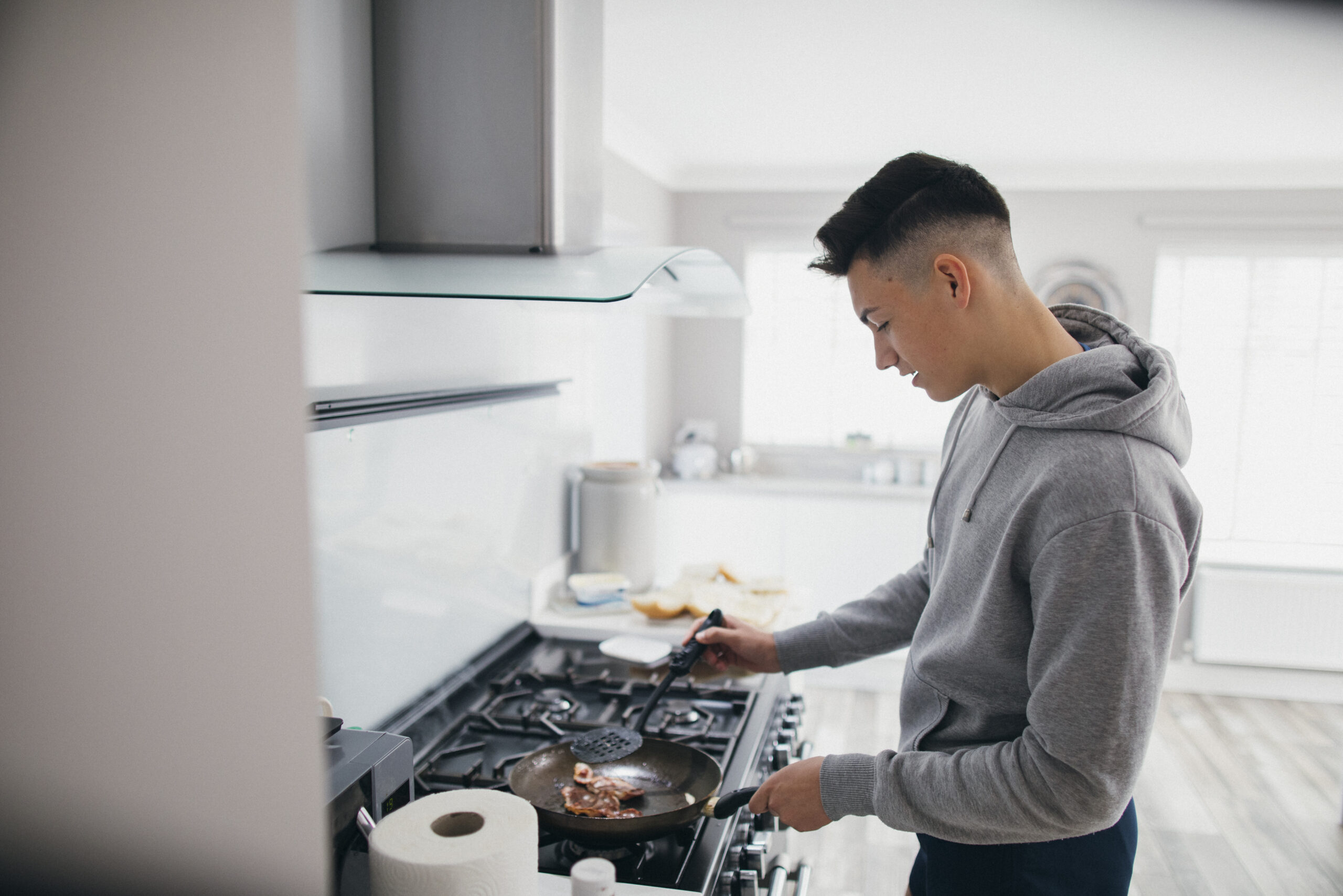A person with short dark hair is cooking bacon in a frying pan on a stove in a modern kitchen. The individual is wearing a gray hoodie and is holding a spatula in their hand while standing in front of the stove. The countertop has a roll of paper towels.